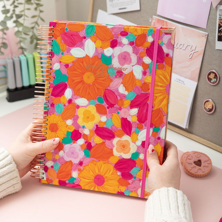 Person holding a colorful floral notebook on a desk with stationery items.