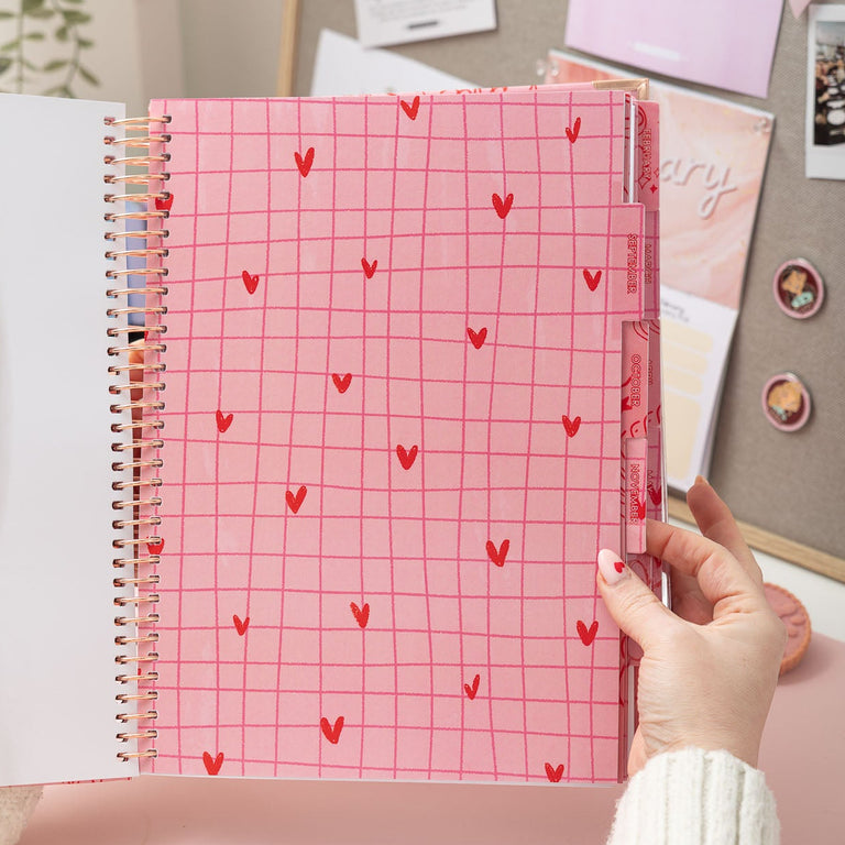 Person holding a pink notebook with heart patterns on a desk with photos and stationery.