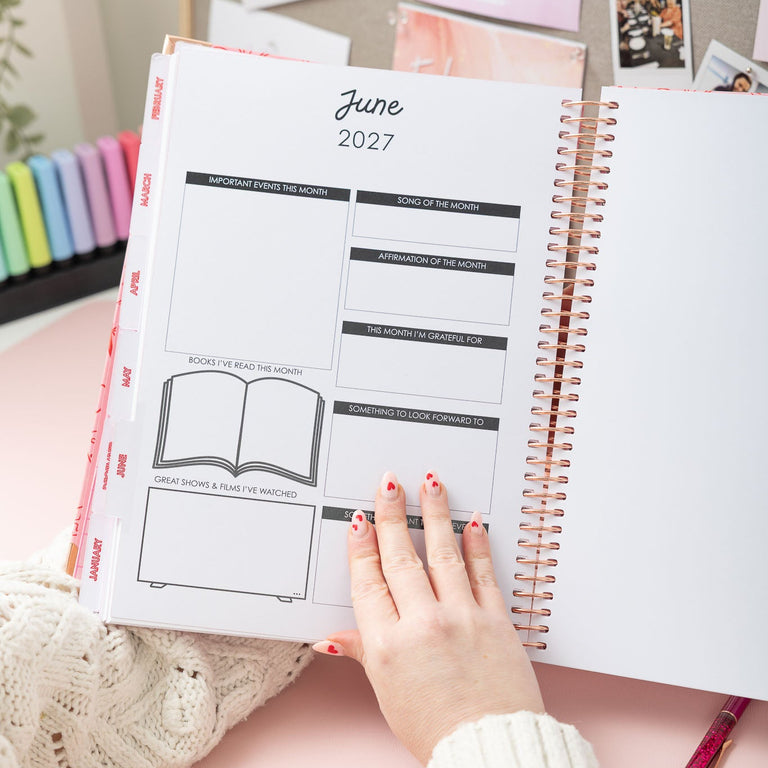 Person holding a planner open to June 2027 with colorful stationery items in the background.