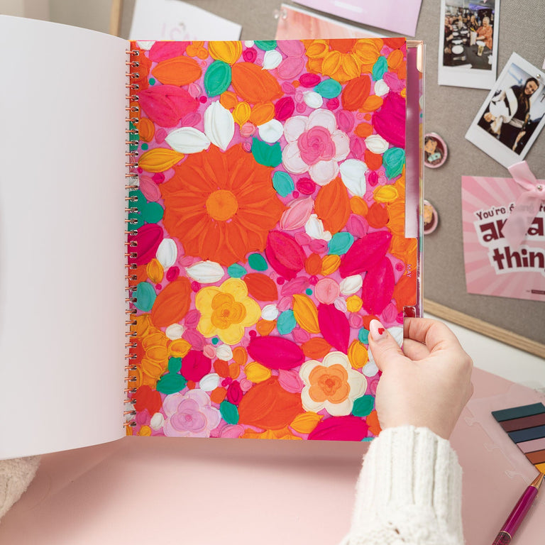 Colorful floral notebook held by a hand on a desk with photos and stationery items.