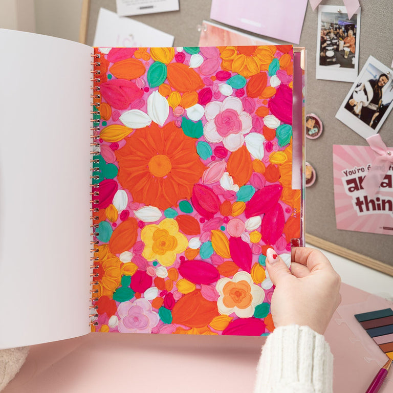 Colorful floral notebook held by a hand on a desk with photos and stationery items.
