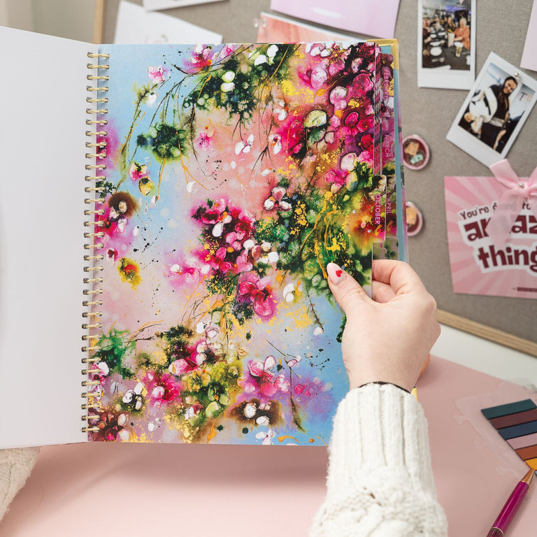 Hand holding a colorful floral notebook against a wall with photos and notes.