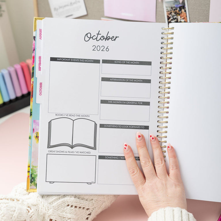 Person holding an open planner showing October 2026, with a pink background and stationery items.
