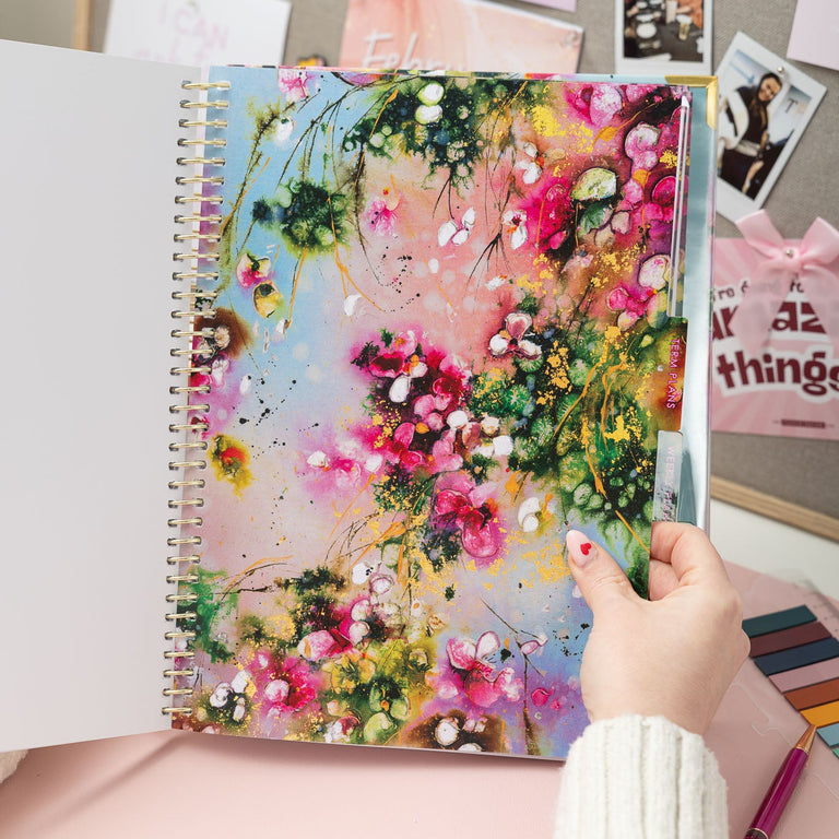 Person holding a spiral-bound notebook with floral design on a desk with photos and stationery.