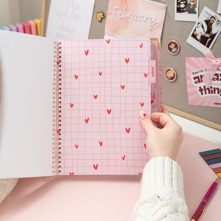 Person holding a pink notebook with heart patterns on a desk with photos and stationery.