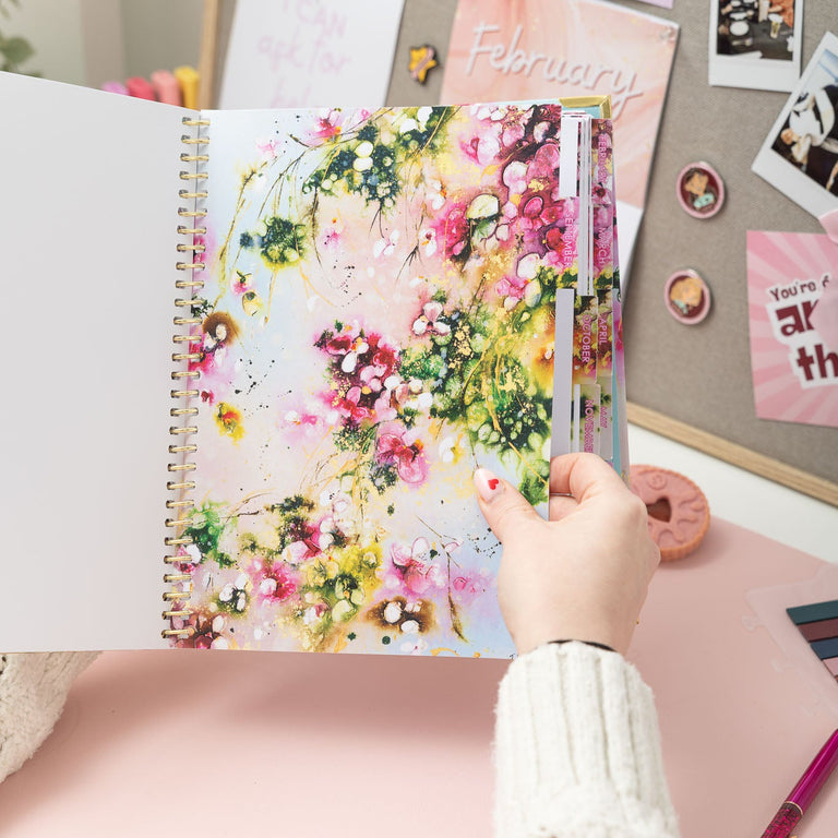 Hand holding a planner with floral design on a desk with decorative items.