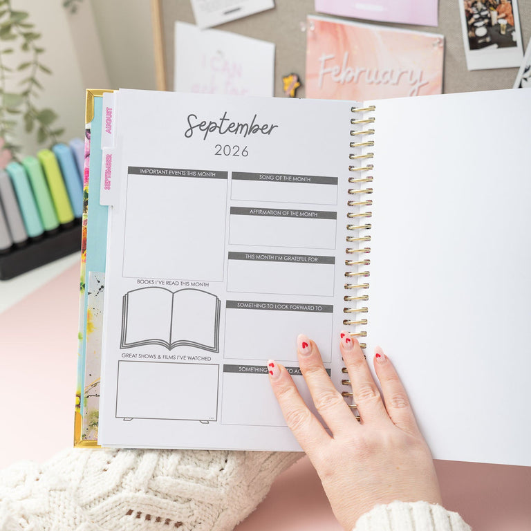 Person holding a planner open to September 2026 on a light pink surface with decorative items in the background.