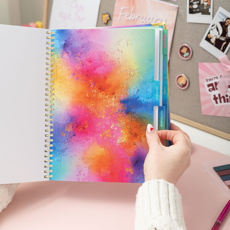 Colorful spiral-bound notebook held by a hand against a background with photos and notes.