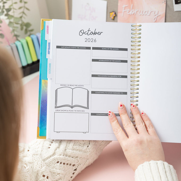 Person holding a planner open to October 2026 with a pink background