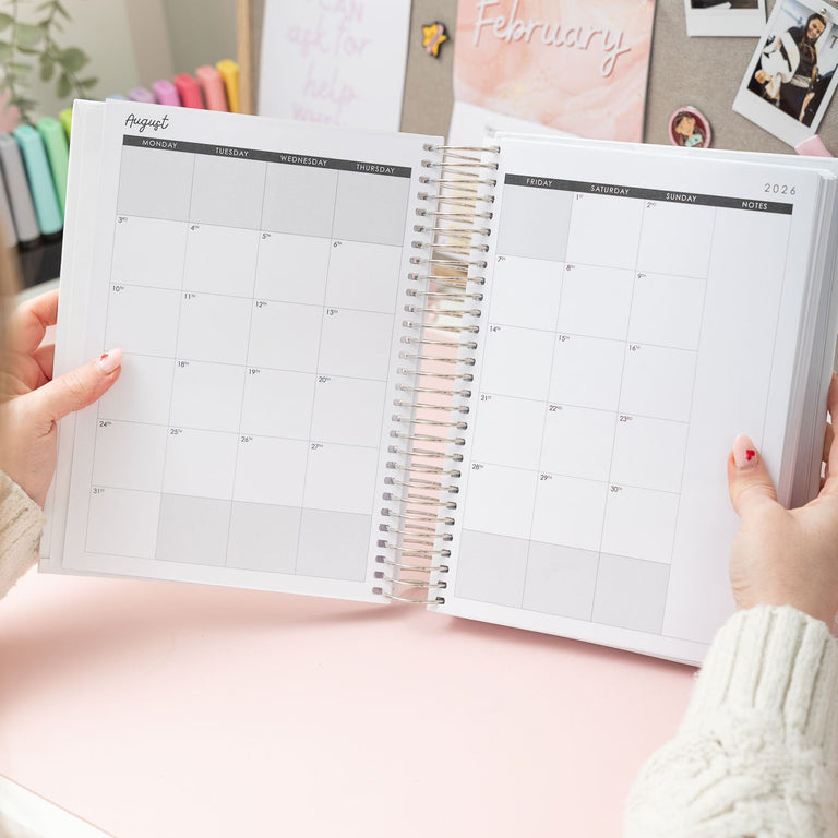 Person holding a spiral-bound planner open on a pink surface with decorative elements.
