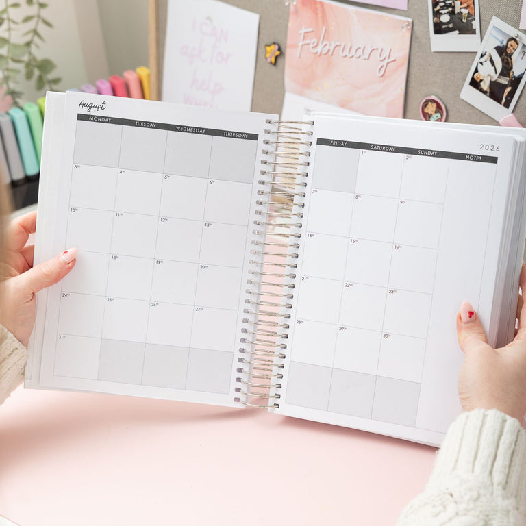 Person holding a spiral-bound planner open on a pink surface with decorative elements.