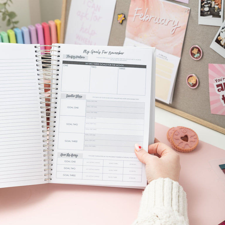 Person holding a planner with a goal-setting page, surrounded by stationery items on a desk.
