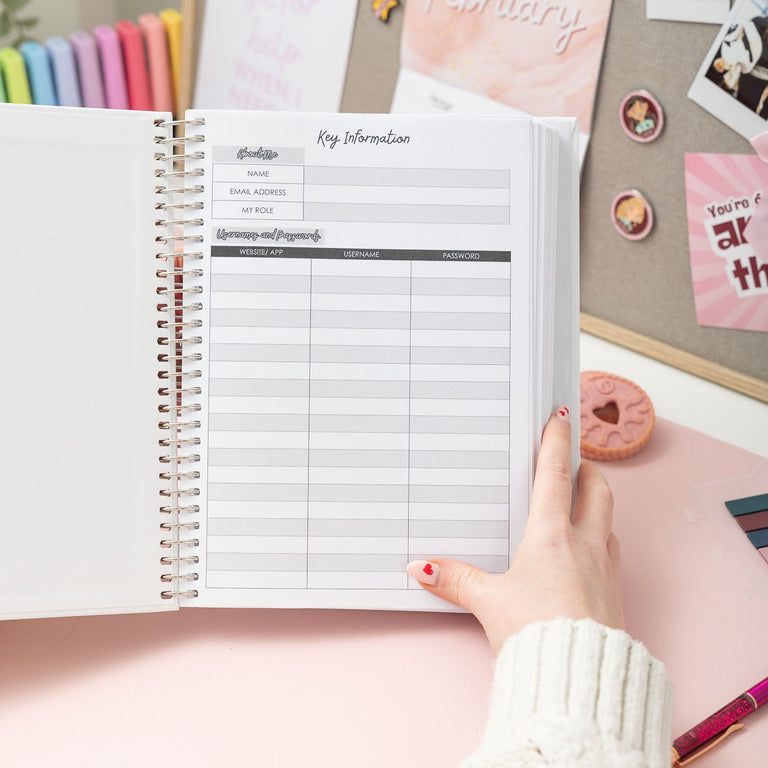 Person holding a spiral-bound planner with colorful pens and decorative items in the background