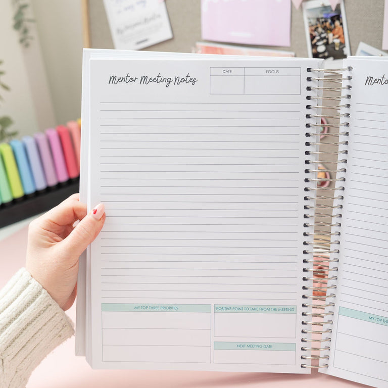 Person holding a spiral-bound notebook with lined pages and a pink background with photos and stationery.