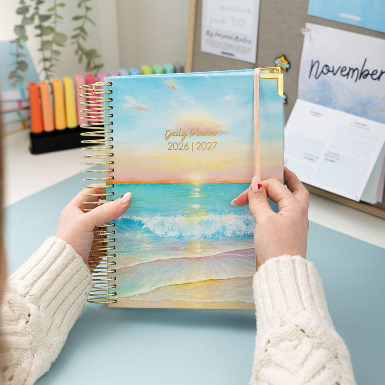 Person holding a spiral-bound planner with a beach design on a desk.