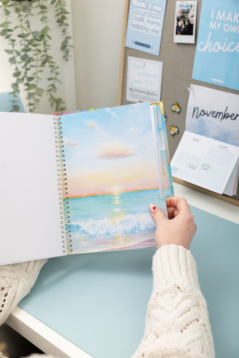 Person holding a planner with a beach scene cover, sitting at a desk.