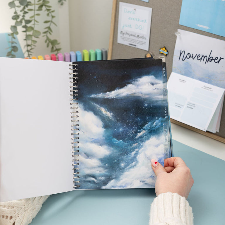 Person holding a spiral-bound notebook with a cloud design against a desk with stationery items.