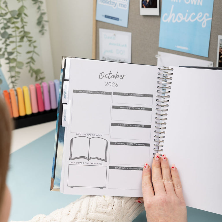 Person holding a planner with October 2023 dates, surrounded by stationery items on a light blue surface.