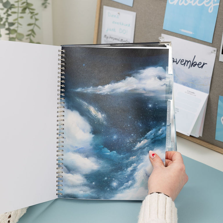 Person holding a spiral-bound notebook with a cloud design on a desk.