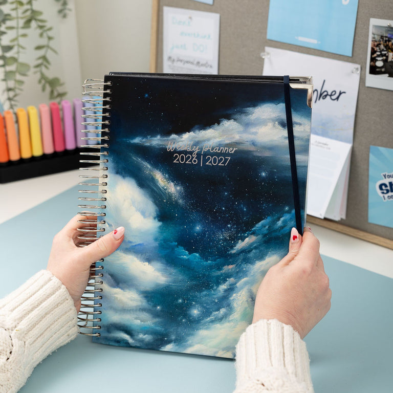 Person holding a planner with a cosmic design on a desk with a bulletin board in the background.