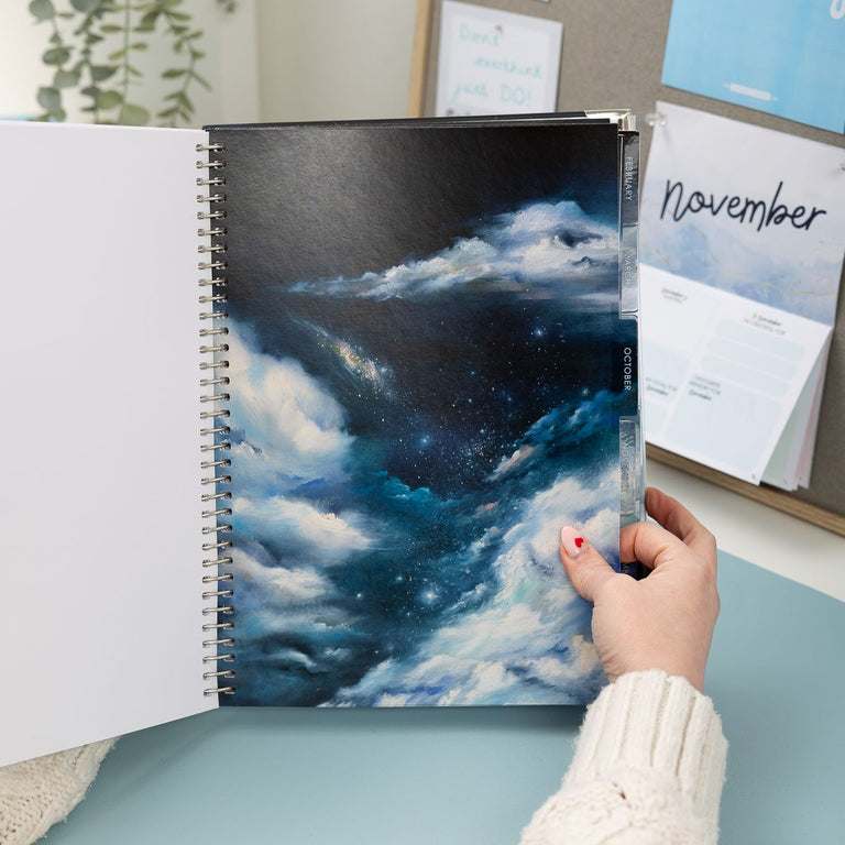 Person holding a spiral-bound notebook with a cloud design, surrounded by office decor.