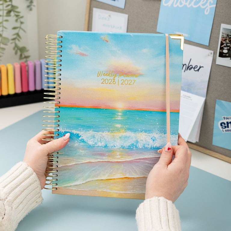 Person holding a planner with a beach design on a desk.