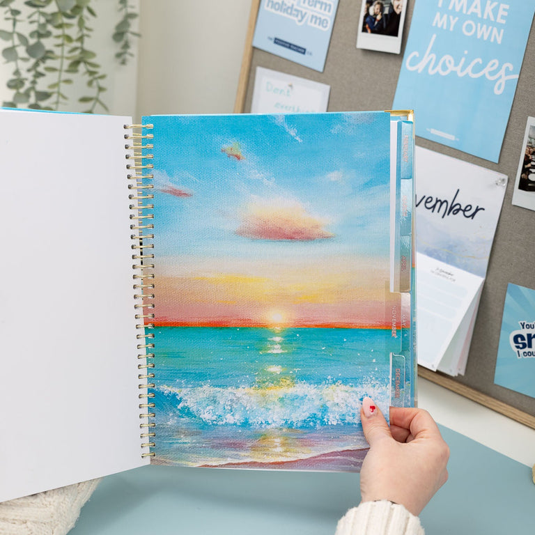 Person holding a spiral-bound notebook with a beach scene cover, placed on a desk with a bulletin board in the background.