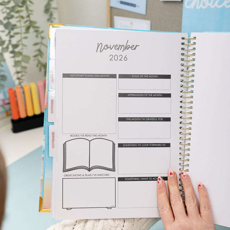 Person holding a planner with November 2026 layout, surrounded by office supplies and decor.