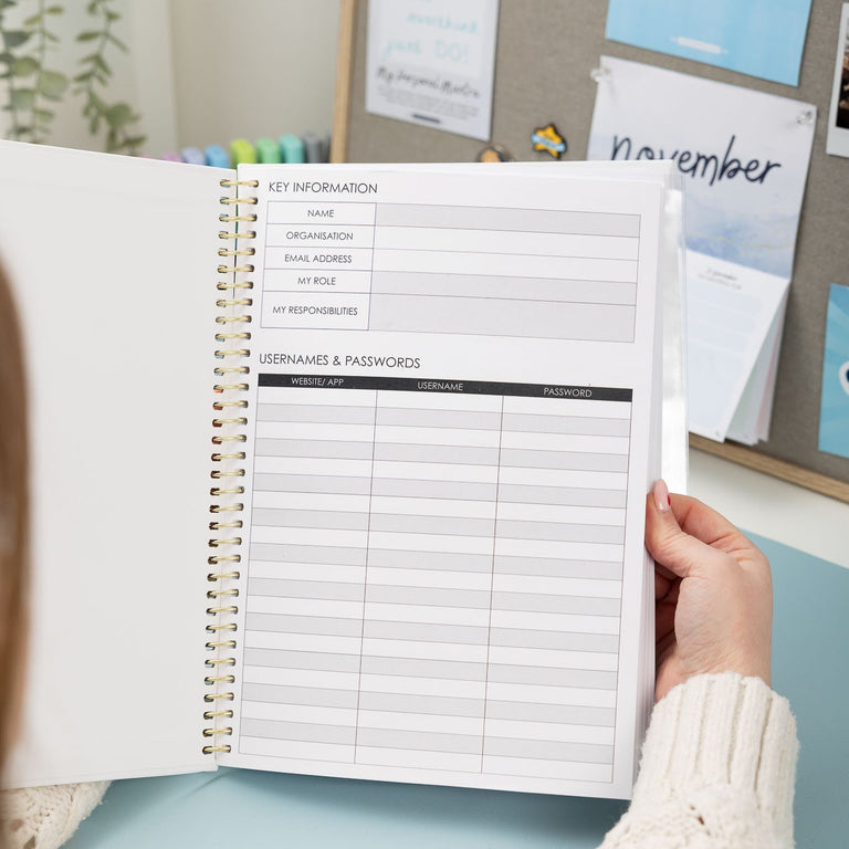 Person holding a planner with a key information and passwords page, in front of a bulletin board.