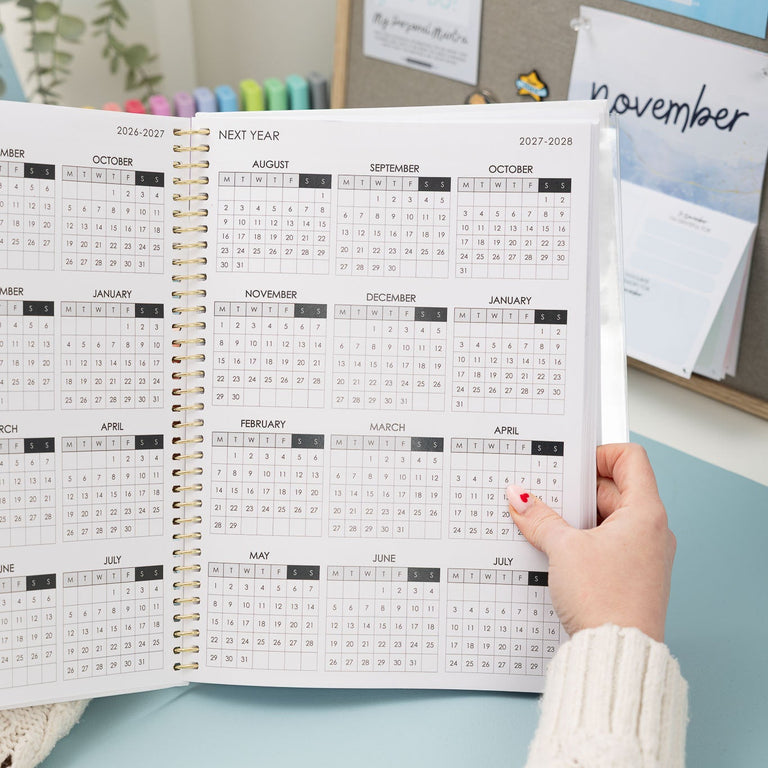 Person holding an open annual planner with a calendar view, sitting at a desk.