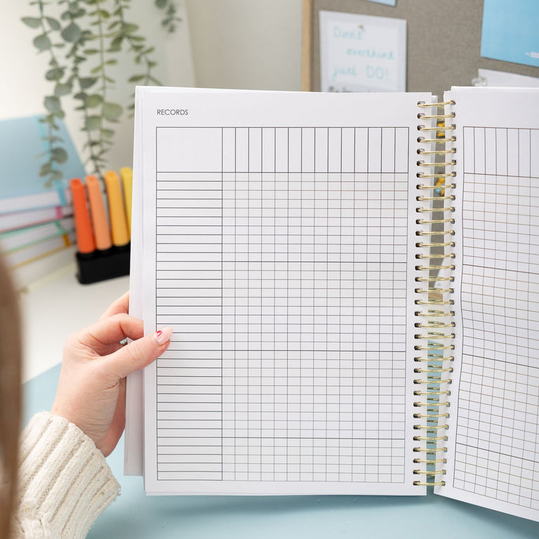 Person holding an open spiral-bound notebook with grid and plain pages, with a light blue background.