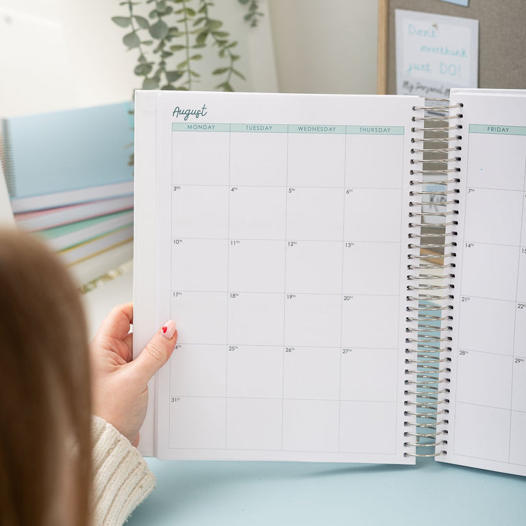 Person holding an open planner with a calendar view, sitting on a light blue chair.