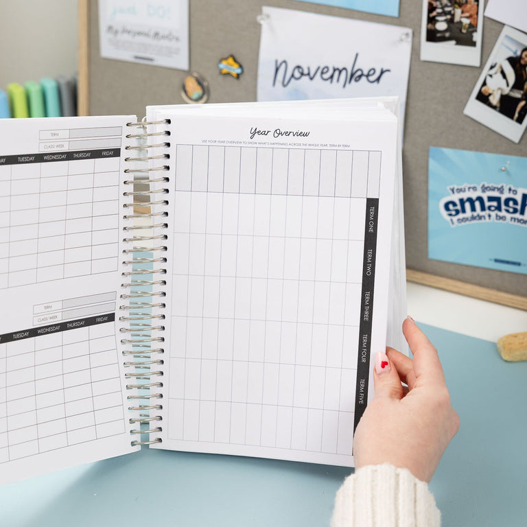 Person holding a planner with a calendar on a desk, surrounded by photos and notes.