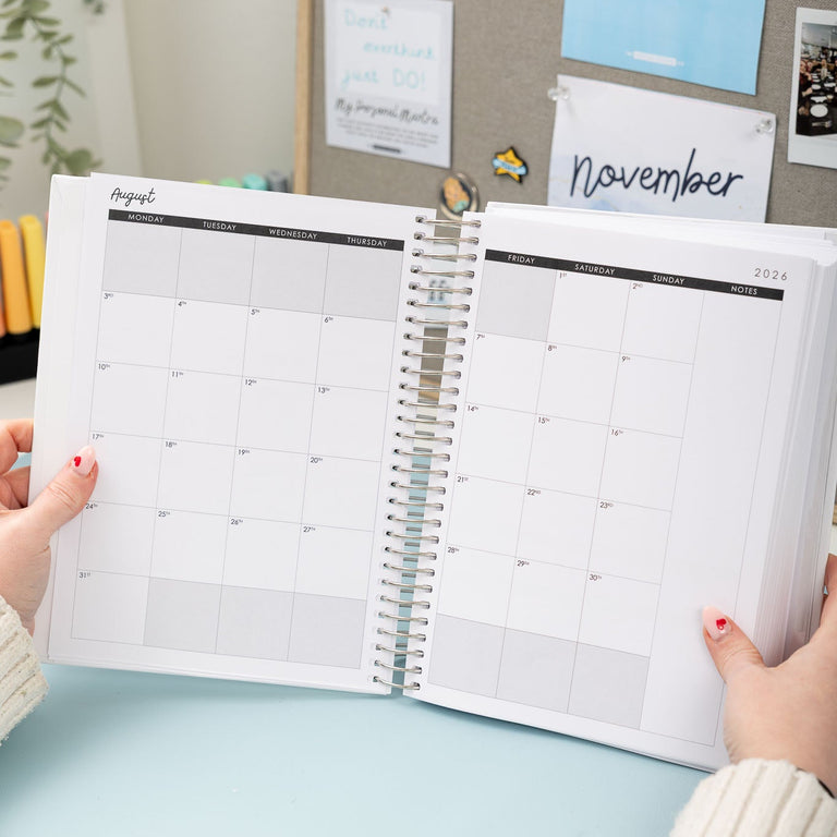 Person holding an open planner with a calendar view, set against a background of a desk with stationery items and wall decor.