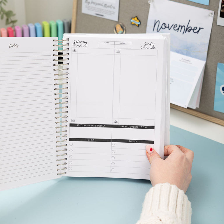 Person holding a planner open on a desk with a bulletin board in the background