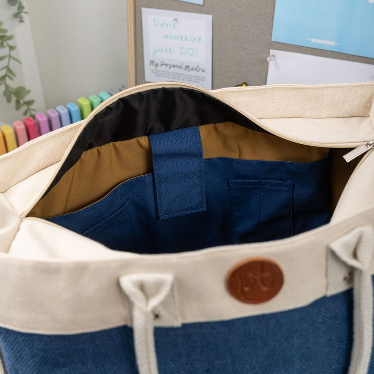 Blue and beige bag with a brown leather patch on a wall with posters and a plant.