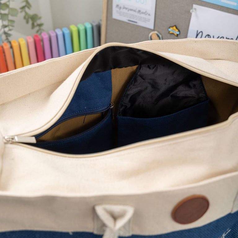 Beige tote bag with a brown button and 'Miss H' text, open to reveal a blue interior, against a background with colorful markers and a bulletin board.