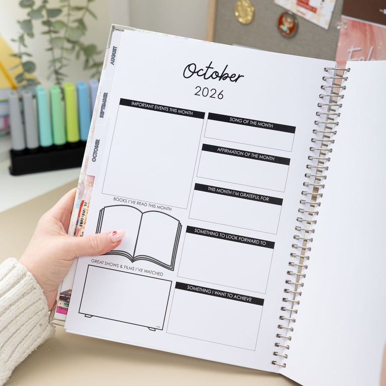 Person holding a planner open to October 2026 on a desk with a decorative background.