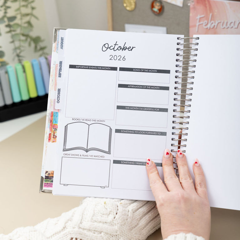 Person holding an open planner with October 2026 dates, sitting at a desk.