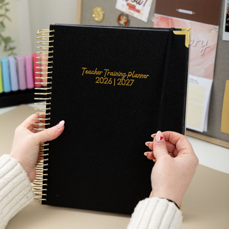 Black planner titled 'Teacher Training Planner 2026-2027' held by a person in a classroom setting.