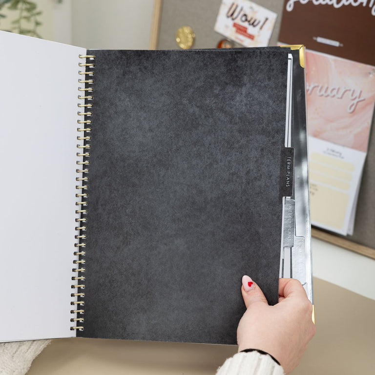 Person holding a spiral-bound notebook with a dark cover in front of a bulletin board.