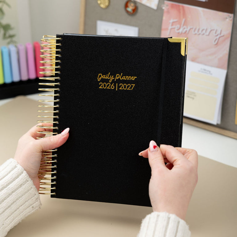 Person holding a black daily planner with gold accents, set against a blurred office background.