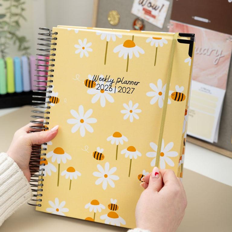 Yellow planner with floral and bee pattern held by a person in an indoor setting.