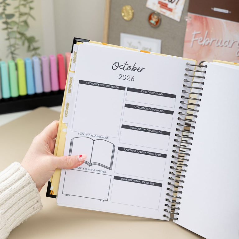Person holding a planner open to October 2026 on a desk with decorative items in the background.