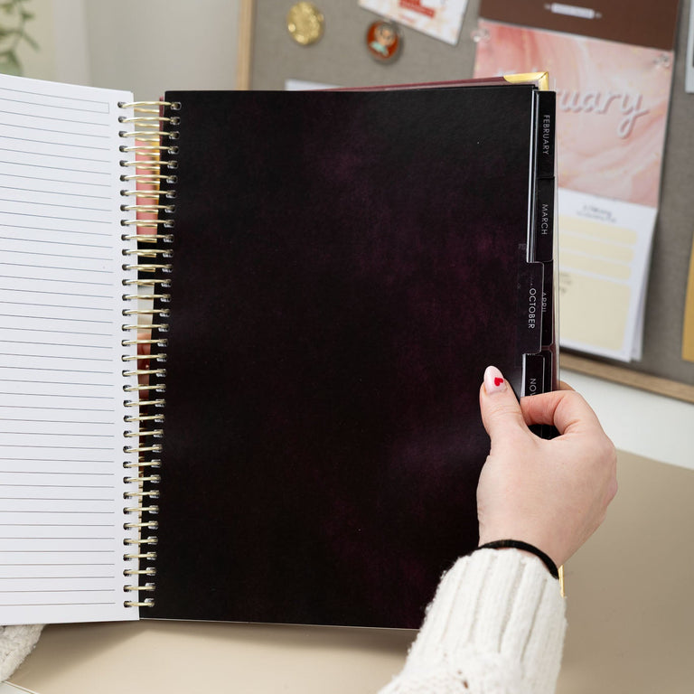 Person holding a black spiral-bound notebook with a white cover on a desk.