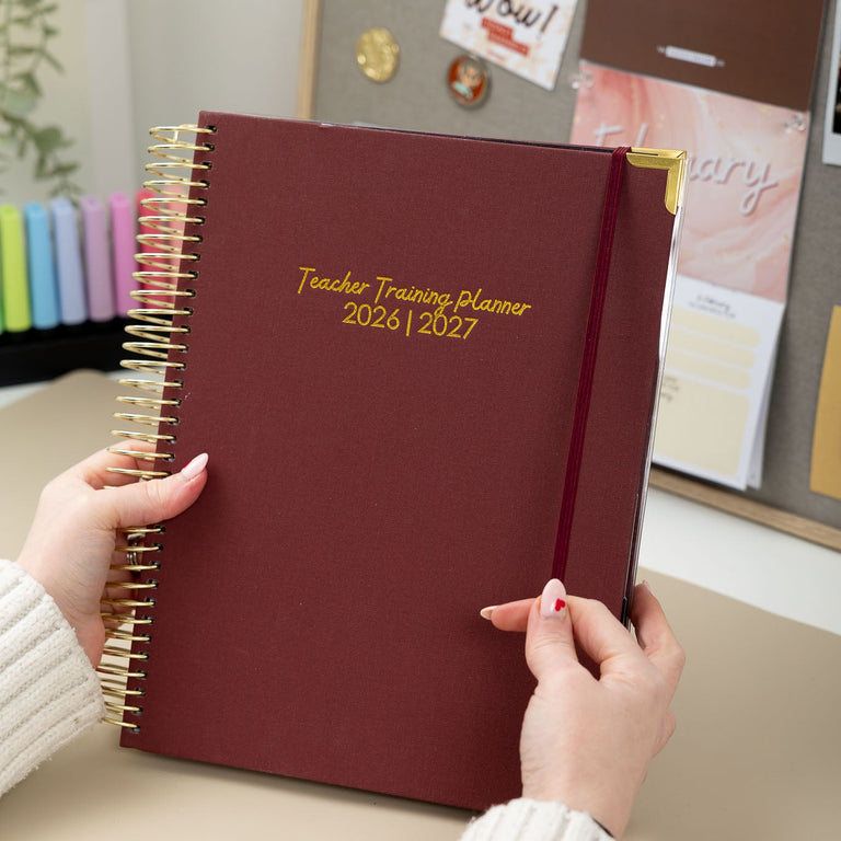 Maroon planner with gold trim held by a person on a desk with a bulletin board in the background.