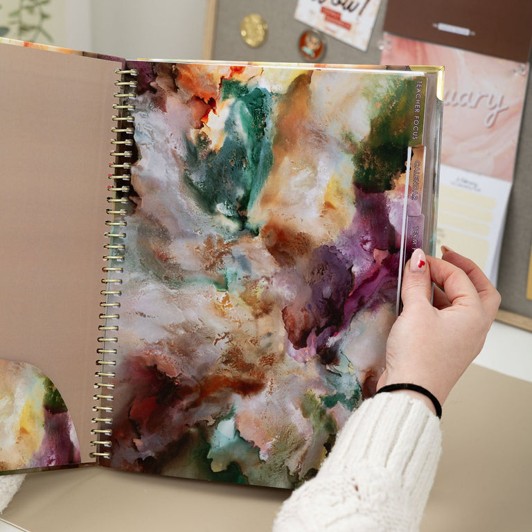 Person holding a spiral-bound notebook with a colorful abstract design on a desk.