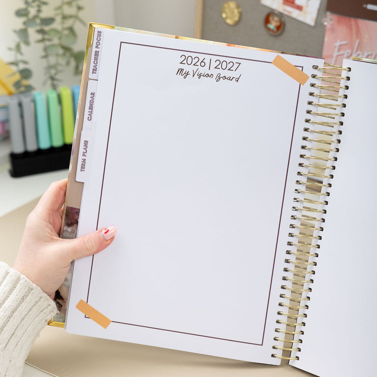 Person holding a spiral-bound planner with 2026-2027 layout on a desk.