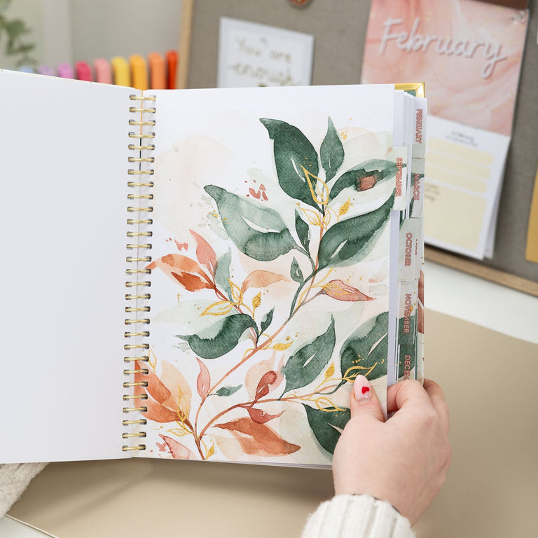 Hand holding a planner with floral design on a desk with stationery items in the background