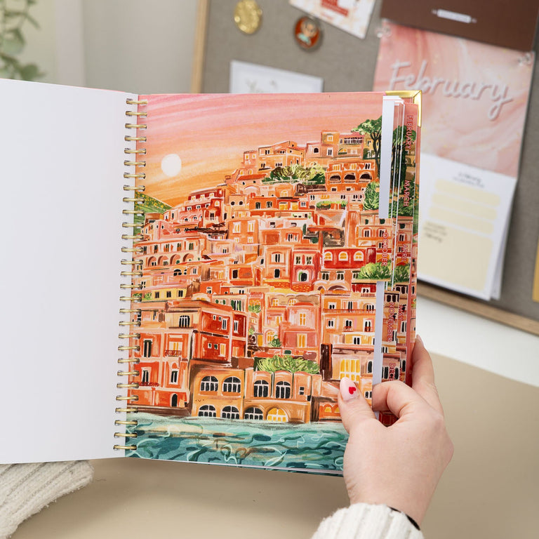 Person holding a spiral-bound notebook with a colorful illustration of a coastal town.